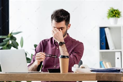 精疲力竭的商人坐在笔记本电脑旁，一边举着眼镜，一边触摸着眼睛 高清摄影大图-千库网
