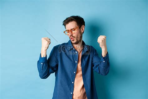 成功的男人戴着新眼镜跳舞并取得胜利，举起拳头手势，说是并庆祝，站在蓝色背景上高清摄影大图-千库网