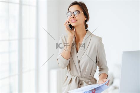 快乐体贴的成功女性员工将现代手机放在耳边，进行电话交谈，看向一边，穿着优雅，审查月度报告，研究图表，站在办公室高清摄影大图-千库网