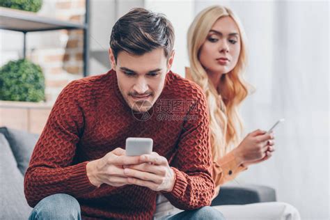 嫉妒的年轻女人用智能手机看着微笑的男朋友在家用智能手机高清图片下载-正版图片503366948-摄图网