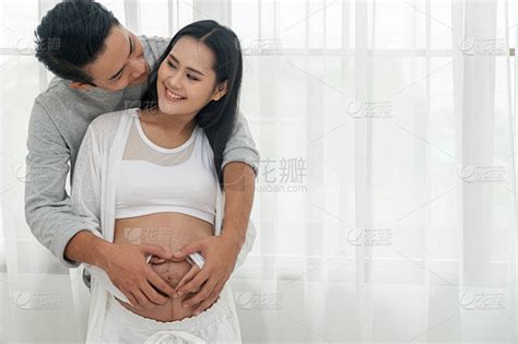 丈夫拥抱妻子，双手在怀孕的肚子上做心形。幸福的一对年轻男子拥抱他的怀孕美丽的妻子，微笑着看着对方的眼