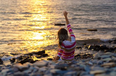 一个女孩坐在海边，把石头扔进水里，举起双手，傍晚，日落高清摄影大图-千库网