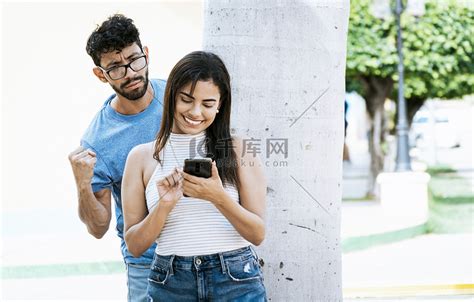 微笑的年轻女子使用智能手机嫉妒的男高清图片下载-正版图片503367028-摄图网