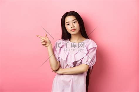 年轻自鸣得意的亚洲女孩看起来很酷，手指留在标志上，在粉红色浪漫背景下做广告产品高清摄影大图-千库网