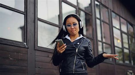 美丽的女孩带着智能手机戴着耳机听音乐，转身跳舞，在城市里散步欣赏歌曲。人与乐的概念。视频素材_ID:VCG42N1048069736-VCG.COM