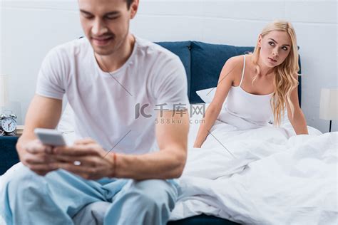 嫉妒的年轻女子看着男友使用智能手机在卧室的前景高清摄影大图-千库网
