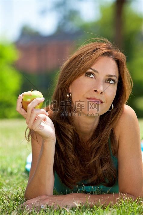 一个三十多岁的漂亮女人躺在外面的草地上，手里拿着或吃着苹果，抬头思考着高清摄影大图-千库网