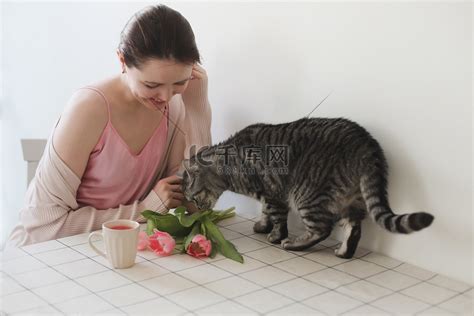 年轻女子和她的猫早上在家里闻到新鲜的粉红色郁金香的味道。高清摄影大图-千库网